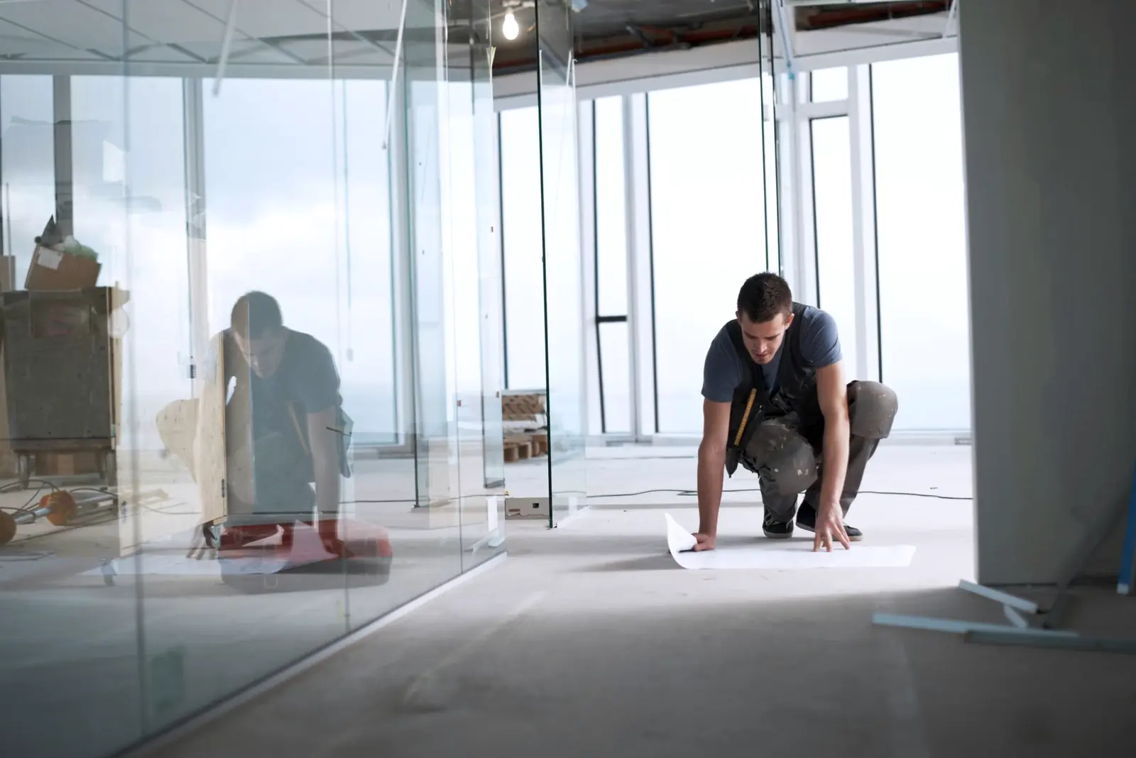 A man kneeling on the floor of a glass-partitioned room while working on a sheet