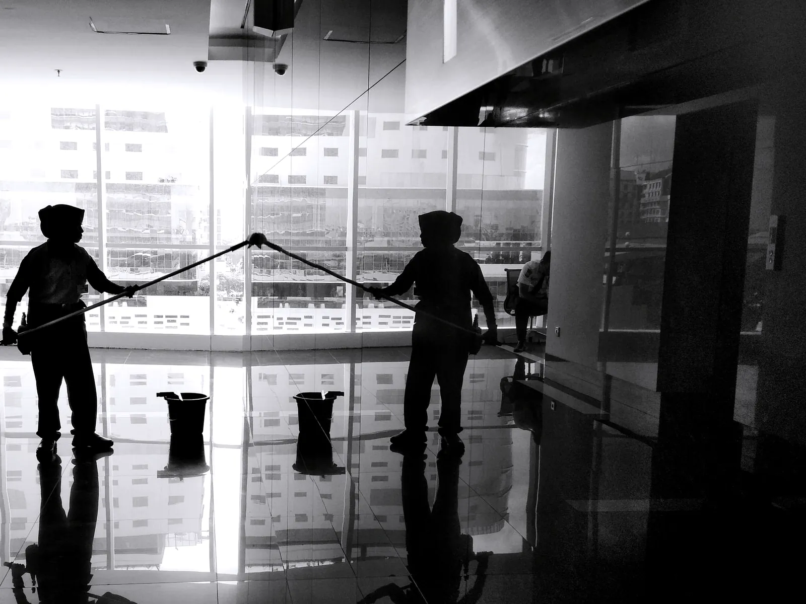 Two cleaners in silhouette working with long poles in front of a tall glass wall, black and white photo